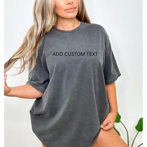 May include: A dark gray, oversized t-shirt with the text "ADD CUSTOM TEXT" printed across the chest. The shirt has short sleeves and a crew neck. The fabric appears to be soft and slightly faded.