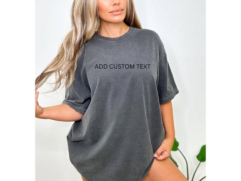 May include: A dark gray, oversized t-shirt with the text "ADD CUSTOM TEXT" printed across the chest. The shirt has short sleeves and a crew neck. The fabric appears to be soft and slightly faded.