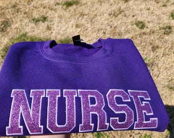 May include: Purple sweatshirt with the word "NURSE" in large, white letters with purple glitter accents.