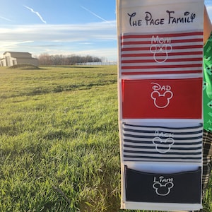 May include: A white fabric organizer with four pockets labeled "Mom", "Dad", "Jack", and "Liam". Each pocket has a red, white, and blue striped fabric with a white Mickey Mouse head embroidered on it. The organizer is hanging on a white wall.