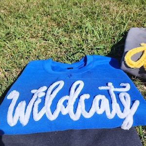 May include: A royal blue sweatshirt with the word "Wildcats" in white, fluffy, cursive lettering. The sweatshirt is folded on top of a black garment. The background is green grass.