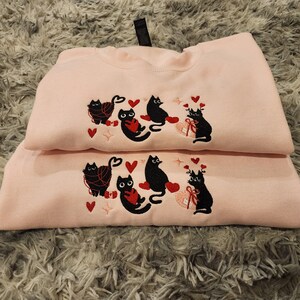 May include: Two pink sweatshirts with black embroidered cats holding red hearts. The cats are wearing yarn around their necks. The sweatshirts are stacked on top of each other.