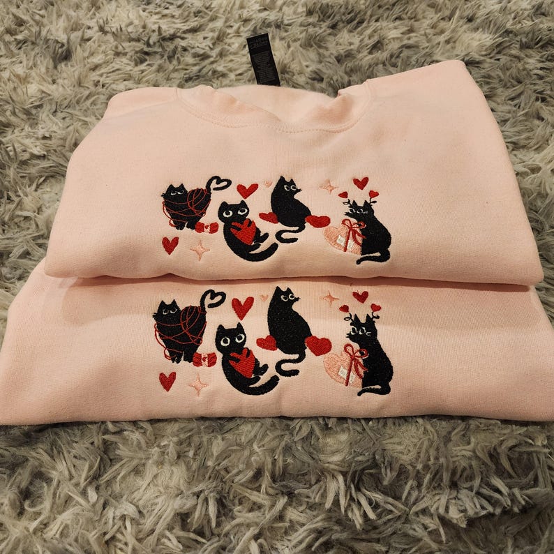 May include: Two pink sweatshirts with black embroidered cats holding red hearts. The cats are wearing yarn around their necks. The sweatshirts are stacked on top of each other.