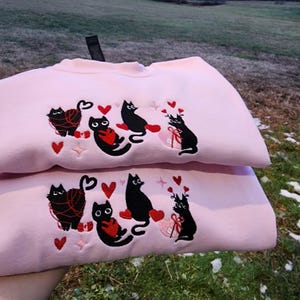 May include: Pink sweatshirt with black embroidered cats holding hearts and yarn. The cats are wearing red hearts and have white eyes. The sweatshirt is folded in half.