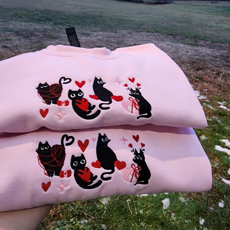 May include: Pink sweatshirt with black embroidered cats holding hearts and yarn. The cats are wearing red hearts and have white eyes. The sweatshirt is folded in half.