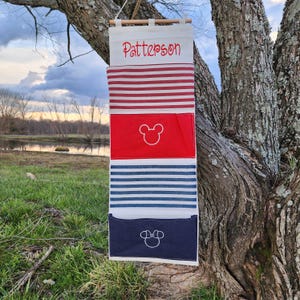 May include: A fabric wall organizer with a wooden dowel and hanging cord. The top section has the name "Patterson" in red script. Below are red and white striped, red with a Mickey Mouse outline, blue and white striped, and navy blue with a Minnie Mouse outline pockets.