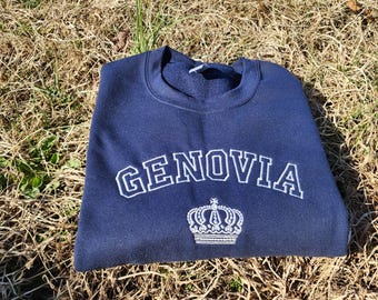 Può includere: Una felpa blu navy con la parola "GENOVIA" ricamata in bianco con una corona bianca sotto.