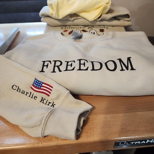 May include: Tan sweatshirts with embroidered text and designs. One sweatshirt reads "FREEDOM" in black. Another has "Gilmore Ghouls" with a ghost and leaf design. A sleeve displays an American flag and the name "Charlie Kirk".