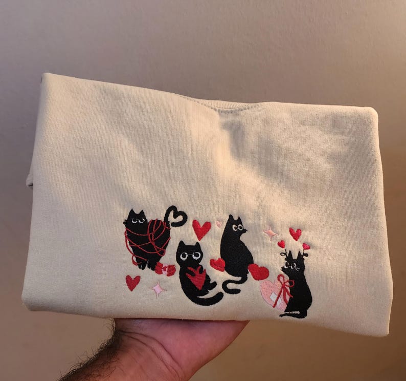 May include: A cream-colored sweatshirt with black embroidered cats and red hearts. The cats are playing with yarn, holding hearts, and wearing a bow.