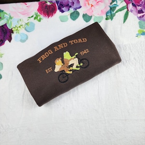 Könnte beinhalten: Ein braunes Sweatshirt mit dem Text "Frog and Toad" und "Est. 1942", das in Orange gestickt ist. Das Design zeigt zwei Frösche, die Fahrrad fahren.