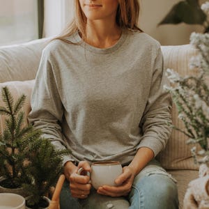 May include: A woman wearing a gray long-sleeved shirt and blue jeans sits on a couch holding a white ceramic mug.  She is surrounded by potted plants.