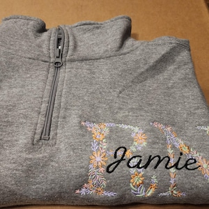 May include: Grey fleece pullover with a zip and floral embroidery that spells out the name "Jamie".