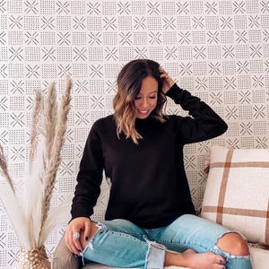 以下が含まれることがあります： 黒い長袖スウェットシャツと、明るい青色の破れたジーンズ。装飾的な枕と白いフェイクファーの毛布が付いたソファに座っている人。背景には、黒い幾何学模様の白い壁があります。