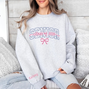 May include: A light grey sweatshirt featuring the text "SENIOR class of 2026" in blue and pink, with a pink bow graphic. The name "Kelsey" is printed in pink on the left sleeve. The sweatshirt is a casual style.