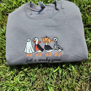 May include: A gray sweatshirt with an embroidered design of four cartoon geese dressed in Halloween costumes. The geese are a ghost, a vampire, a jack-o'-lantern, and a skeleton. The text "just a spooky goose" is embroidered below the geese.