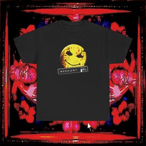 Puede incluir: Camiseta negra con un diseño de cara sonriente amarilla y salpicaduras de sangre. La palabra "MANHUNT" está impresa en blanco en una etiqueta rectangular debajo del gráfico. La camiseta tiene cuello redondo y mangas cortas.