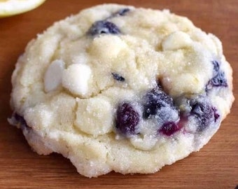 May include: A close-up of a blueberry and white chocolate chip cookie. The cookie has a light-colored, slightly textured exterior with visible blueberries and white chocolate chips. The cookie is on a wooden surface.