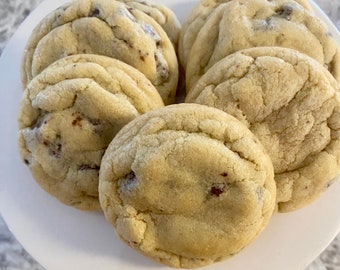 May include: A collection of six round, golden-brown chocolate chip cookies arranged on a white plate. Each cookie features a soft, textured surface with visible dark chocolate chips embedded throughout the baked dough.