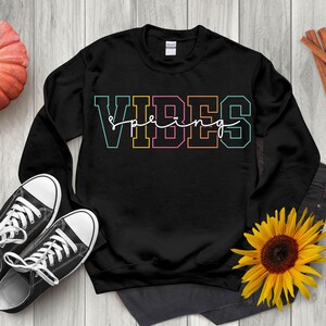 Puede incluir: Sudadera negra con las palabras "VIBES Spring" en una fuente colorida de estilo universitario. La sudadera se muestra con zapatillas negras, un girasol y unos vaqueros.