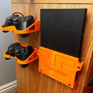 Puede incluir: Soporte de pared naranja para una consola PlayStation 2 y dos mandos de videojuegos negros. El soporte de la consola tiene el texto "PlayStation 2" impreso en él.