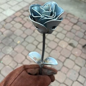 May include: A handcrafted metal rose sculpture with a stem and two leaves. The rose petals are layered and detailed, with a silver-blue hue. The sculpture is held by a person wearing a brown glove. The background is a brick patio.