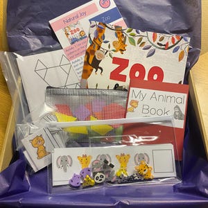 May include: A purple and white gift box filled with a variety of educational toys for children. The box contains a book titled "My Animal Book", a clear plastic bag with colorful shapes, a small mesh bag with animal-themed buttons, and a worksheet with a geometric shape.