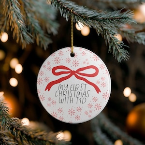 May include: A round white Christmas ornament with a red bow and snowflakes. The text "MY FIRST CHRISTMAS WITH T1D" is written in black script. The ornament hangs from a gold string against a blurred Christmas tree background.