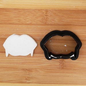 May include: A white plastic vampire teeth cookie cutter and a black plastic vampire teeth cookie cutter on a wooden surface.