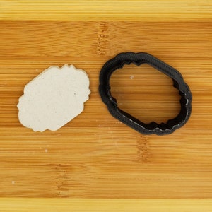 May include: A white clay cookie dough cutout in the shape of a decorative oval frame, next to a black plastic cookie cutter in the same shape.