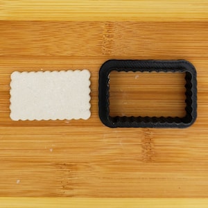 May include: A white cookie with a scalloped edge sits next to a black plastic cookie cutter with a scalloped edge on a wooden surface.