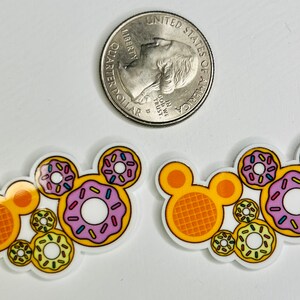 May include: Two sets of Mickey Mouse ears made of resin with a donut theme. Each set features a waffle Mickey Mouse ear with a yellow and orange waffle pattern and a donut ear with pink, yellow, and white donuts with sprinkles.