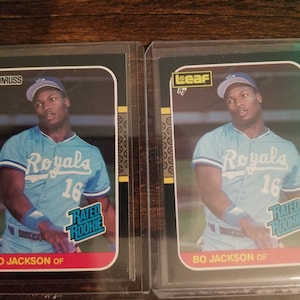May include: Two baseball cards featuring Bo Jackson, a former professional baseball player. Both cards are black with gold accents and feature a photo of Jackson in a Kansas City Royals uniform. The card on the left is a Donruss card from 1987, and the card on the right is a Leaf card from 1987.