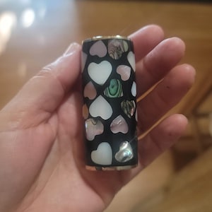 Può includere: Un tubo di rossetto nero ornato da intarsi a forma di cuore di conchiglia di abalone bianca, rosa e iridescente. I bordi superiore e inferiore sono rifiniti con oro. Il design è un mix di eleganza e fantasia.