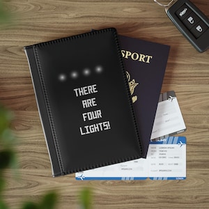 以下が含まれることがあります： 黒いパスポートホルダーに「THERE ARE FOUR LIGHTS!」という白い文字が印刷されています。ホルダーはパスポートと航空券の上に置かれています。