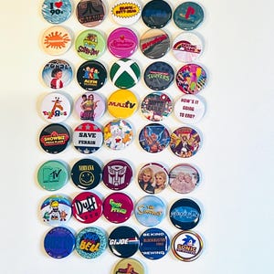Puede incluir: Una colección de botones redondos con diseños de los años 90. Los botones presentan logotipos y personajes de programas de televisión, películas y dibujos animados populares. Los botones son de varios colores y tamaños, con texto y gráficos.
