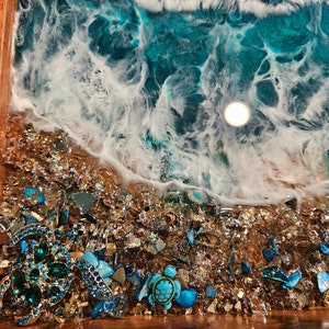 May include: A wooden tray featuring a resin ocean scene. The resin is blue and white, with a wave breaking on a beach of gold glitter and blue and green glass pieces. There are two jeweled turtle figurines on the beach.