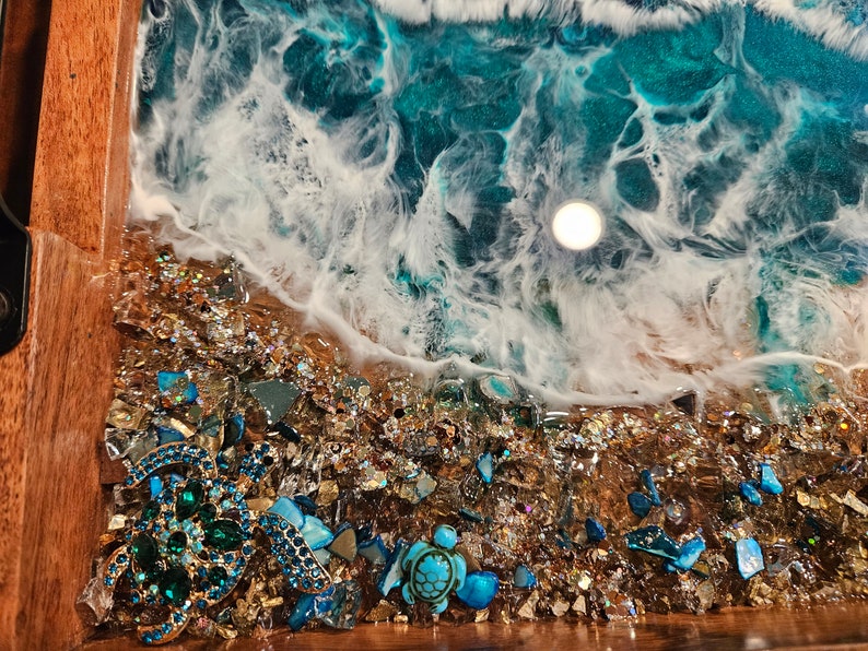 May include: A wooden tray featuring a resin ocean scene. The resin is blue and white, with a wave breaking on a beach of gold glitter and blue and green glass pieces. There are two jeweled turtle figurines on the beach.