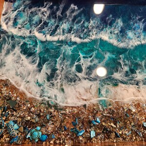May include: A close-up of a resin art tray featuring a blue and white ocean wave design. The tray has a wooden frame and a layer of gold glitter and blue and green glass pieces at the bottom.
