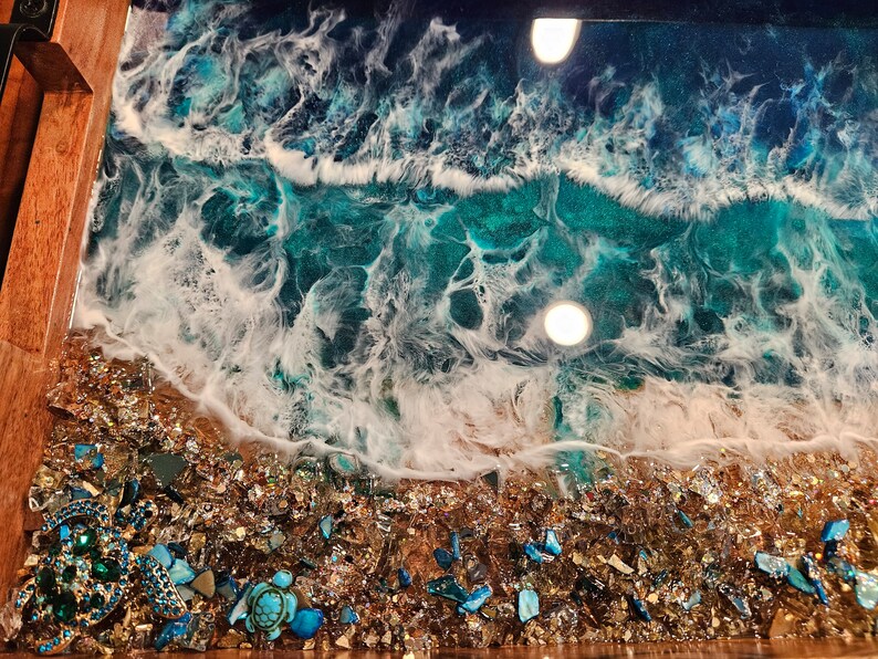 May include: A close-up of a resin art tray featuring a blue and white ocean wave design. The tray has a wooden frame and a layer of gold glitter and blue and green glass pieces at the bottom.