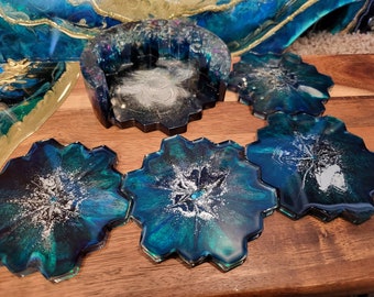 Puede incluir: Conjunto de seis posavasos hexagonales con base de resina azul verdoso, azul y negro con detalles de purpurina plateada. Los posavasos están dispuestos en semicírculo sobre una superficie de madera.