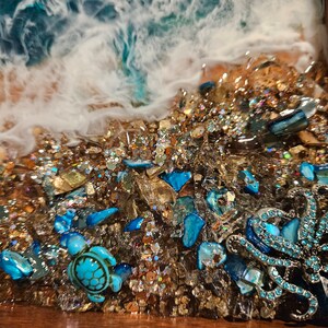 May include: Close-up of a resin art piece depicting a beach scene with a blue octopus and a sea turtle. The piece is filled with gold glitter and blue and gold glass pieces.