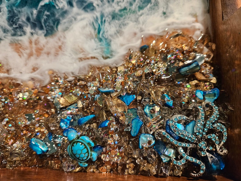 May include: Close-up of a resin art piece depicting a beach scene with a blue octopus and a sea turtle. The piece is filled with gold glitter and blue and gold glass pieces.