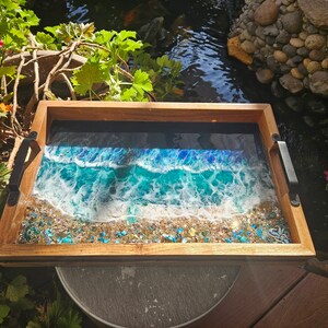 May include: A wooden serving tray with a resin inlay depicting a blue ocean wave crashing on a sandy beach. The tray has black metal handles.