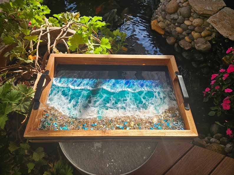 May include: A wooden serving tray with a resin inlay depicting a blue ocean wave crashing on a sandy beach. The tray has black metal handles.