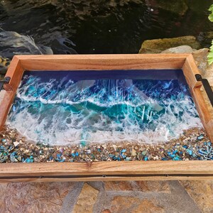 May include: A wooden serving tray with a resin inlay depicting an ocean wave crashing on a sandy beach. The resin is clear and shows blue, green, and white colours. The tray has black metal handles on each side.