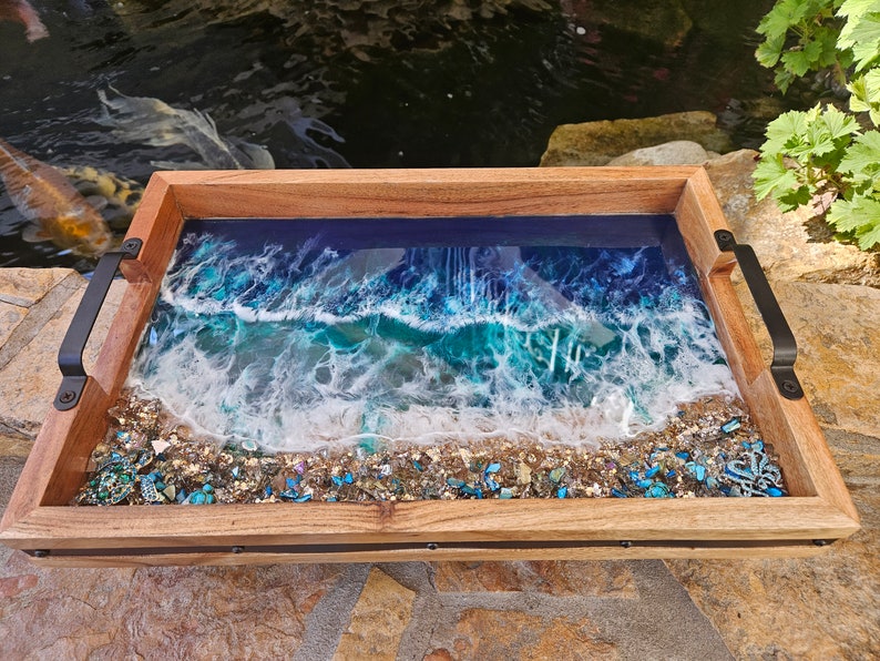 May include: A wooden serving tray with a resin inlay depicting an ocean wave crashing on a sandy beach. The resin is clear and shows blue, green, and white colours. The tray has black metal handles on each side.