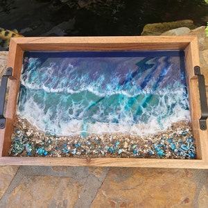 May include: A wooden serving tray with black handles features a resin inlay depicting a blue ocean wave crashing on a sandy beach with turquoise and white accents.
