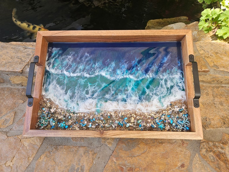 May include: A wooden serving tray with black handles features a resin inlay depicting a blue ocean wave crashing on a sandy beach with turquoise and white accents.