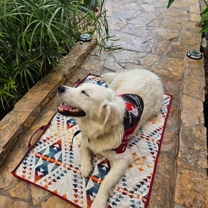 May include: A white dog with a red bandana that says "Service Dog" is lying on a colorful patterned dog bed. The dog is looking up and has its mouth slightly open.