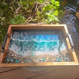 May include: A wooden serving tray with black metal handles features a resin ocean wave design with blue, green, and white colours. The tray is filled with small, iridescent blue and gold glitter.